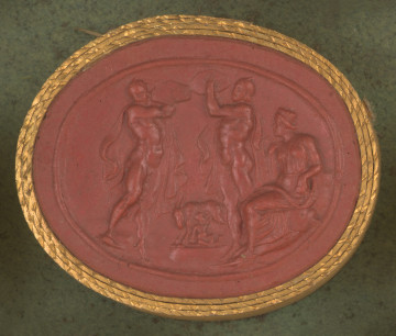 czerwona owalna gemma w grubym złotym obramowaniu; widoczne trzy postaci, dwóch magich mężczyzn w pelerynach - kapłanów - stoi nad kozą (Almateją), która karmi małe dziecko (Zeusa). Kapłani uderzają o tarcze, które trzymają na wysokości swoich twarzy; po prawej stronie na kamieniu siedzi kobieta (Rea, matka Zeusa). 